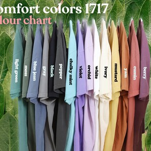 May include: A color chart of Comfort Colors 1717 t-shirts, showcasing a variety of hues. The shirts are displayed in a row, with colors including light green, moss, blue jean, gray, black, pepper, chalky mint, violet, orchid, white, ivory, mustard, yam, cumin, and berry.