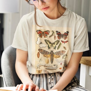 Op de afbeelding: Een wit T-shirt met een vintage illustratie van verschillende vlinders en motten. De illustratie is in tinten bruin, geel, oranje en zwart.