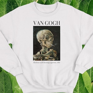 Puede incluir: Sudadera blanca con una impresión negra de una pintura de Van Gogh. La pintura representa un esqueleto con un cigarrillo encendido. El texto debajo de la pintura dice "Skeleton with burning cigarette, 1886".