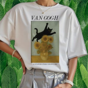 以下が含まれることがあります： 白いTシャツ。ゴッホの「ひまわり」の絵のグラフィックで、黒猫がひまわりの上を跳んでいます。「VAN GOGH」の文字が画像の上にあり、「SUNFLOWERS (THIRD VERSION), 1888」が下にあります。