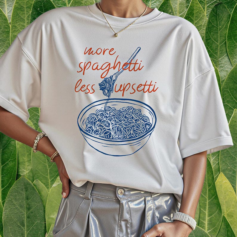 Puede incluir: Camiseta blanca con un dise&ntilde;o azul y rojo. El dise&ntilde;o muestra un cuenco de espaguetis con un tenedor y el texto "more spaghetti less upsetti". La camiseta tiene mangas cortas.