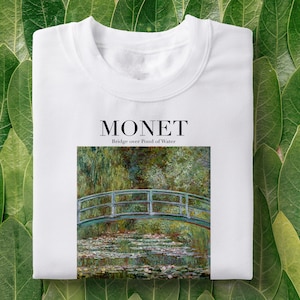 Può includere: Maglietta bianca con una stampa del dipinto di Claude Monet "Ponte sul laghetto con ninfee" e il testo "MONET Ponte sul laghetto con ninfee".