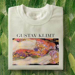 Pode incluir: Uma camiseta branca com uma estampa da pintura "O Beijo" de Gustav Klimt. A pintura representa duas figuras se abraçando, com um fundo de padrões dourados e florais.