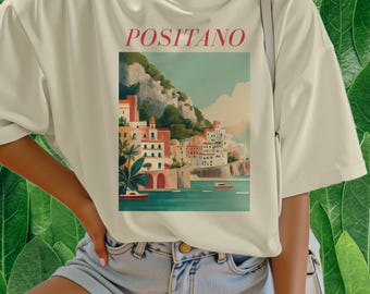 May include: Cream-colored t-shirt featuring a scenic illustration of Positano, Italy, with colorful buildings, boats, and a mountain backdrop. The word "POSITANO" is printed in red above the image. The shirt is made of a soft material.