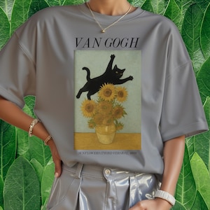 以下が含まれることがあります： 黒猫がひまわりの花瓶の上を跳ねている灰色のTシャツ。ゴッホの「ひまわり」のパロディ。画像の上には「VAN GOGH」、下には「SUNFLOWERS (THIRD VERSION), 1888」と書かれています。