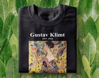 May include: Black t-shirt with a white print of a painting by Gustav Klimt. The painting is of a woman with long dark hair and a colorful dress. The text "Gustav Klimt 1917-1918" is printed below the painting.