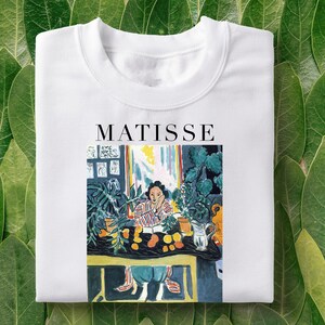 Pode incluir: Camiseta branca com uma pintura colorida de uma mulher sentada a uma mesa com plantas e frutas. O texto "MATISSE" está impresso acima da pintura.