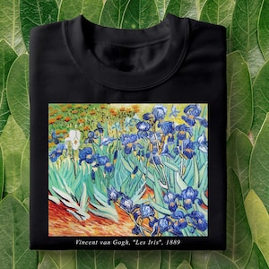Pode incluir: Camiseta preta com uma estampa da pintura "Les Iris" de Vincent van Gogh. A pintura apresenta um campo de íris azuis com folhas verdes e um fundo amarelo. O texto "Vincent van Gogh, "Les Iris", 1889" está impresso abaixo da pintura.