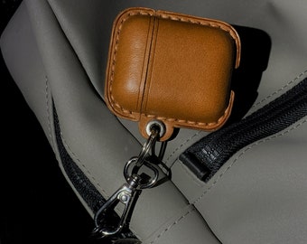 Puede incluir: Una funda de cuero marrón para AirPods con un mosquetón y un llavero plateados, sujeta a una bolsa gris. La funda tiene un borde cosido y líneas verticales en el centro. La bolsa tiene una cremallera negra.