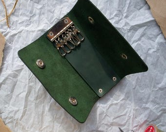May include: A green leather key holder with a metal key ring and five key loops. The holder is open and the inside is lined with a dark green fabric.