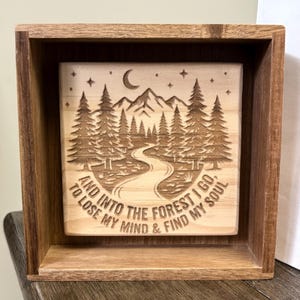 May include: A square wooden shadow box frame containing a light-colored wooden plaque with a laser-engraved forest scene. The design features mountains, pine trees, a crescent moon, stars, a winding path, and the text: AND INTO THE FOREST I GO, TO LOSE MY MIND & FIND MY SOUL.