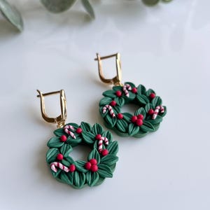 May include: A pair of green Christmas wreath earrings with red berries and white candy canes. The earrings are made of polymer clay and have gold hooks.