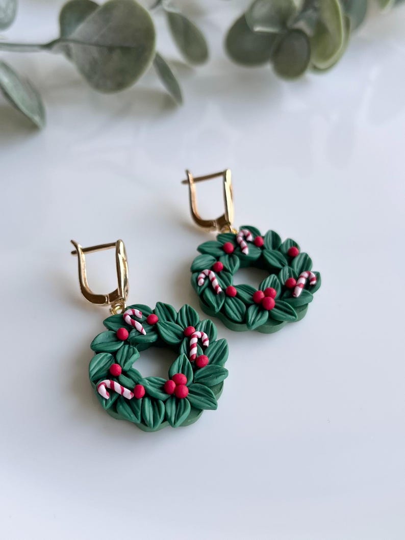 May include: A pair of green Christmas wreath earrings with red berries and white candy canes. The earrings are made of polymer clay and have gold hooks.