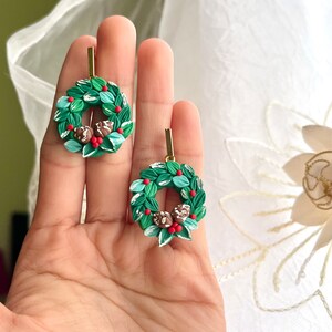 May include: A pair of green and white Christmas wreath earrings with red berries and brown pinecones. The earrings are made of polymer clay and have gold hooks.