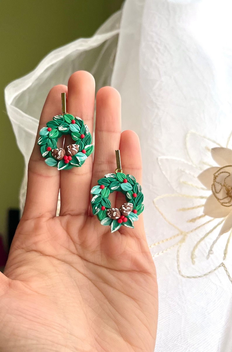 May include: A pair of green and white Christmas wreath earrings with red berries and brown pinecones. The earrings are made of polymer clay and have gold hooks.