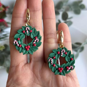 May include: A pair of green Christmas wreath earrings with red berries and white candy canes. The earrings are made of polymer clay and have gold hooks.