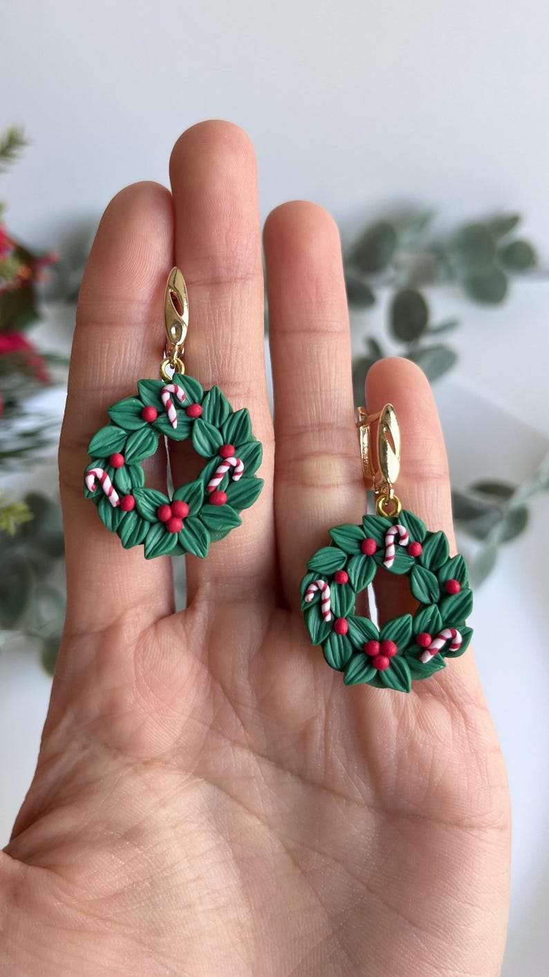 May include: A pair of green Christmas wreath earrings with red berries and white candy canes. The earrings are made of polymer clay and have gold hooks.