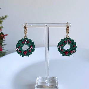 May include: A pair of gold hoop earrings with green and red Christmas wreath charms. The wreaths are decorated with red berries and white candy canes.