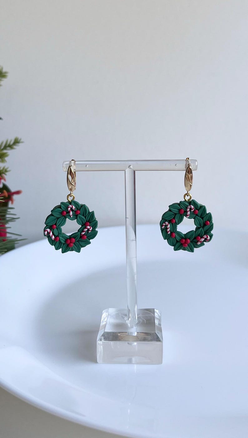 May include: A pair of gold hoop earrings with green and red Christmas wreath charms. The wreaths are decorated with red berries and white candy canes.