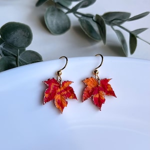 Puede incluir: Un par de pendientes de aro dorados con abalorios de hojas de arce rojas y naranjas. Las hojas tienen un aspecto texturizado y pintado.