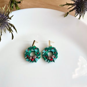 May include: A pair of green and red Christmas wreath dangle earrings with gold hooks. The wreaths are made of polymer clay and feature small red berries and brown pinecones.