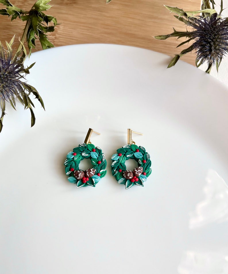 May include: A pair of green and red Christmas wreath dangle earrings with gold hooks. The wreaths are made of polymer clay and feature small red berries and brown pinecones.