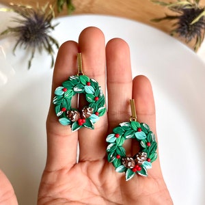 May include: A pair of green and white Christmas wreath earrings with red berries and brown pine cones. The earrings are made of polymer clay and have gold hooks.