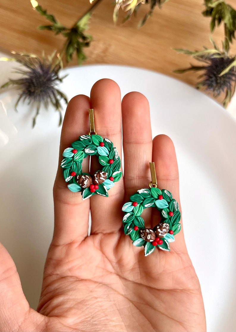 May include: A pair of green and white Christmas wreath earrings with red berries and brown pine cones. The earrings are made of polymer clay and have gold hooks.