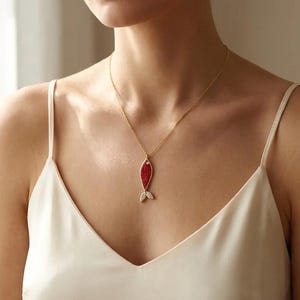 Puede incluir: Collar dorado con un colgante de pez rojo. El colgante de pez tiene un cuerpo rojo texturizado y un pequeño acento de diamante. Las aletas de la cola están adornadas con pequeños cristales transparentes. El collar lo lleva una persona que lleva una camiseta sin mangas blanca.