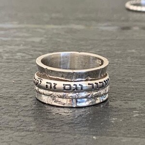 May include: A silver ring with three bands, each with Hebrew text inscribed on it. The ring is on a gray surface.