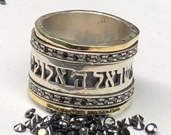 May include: A silver and gold ring with black diamonds and Hebrew writing. The ring is on a white surface with black diamonds scattered around it.