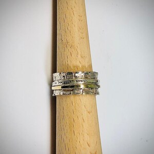 May include: A set of three silver rings stacked on a wooden dowel. The top ring has a textured design, the middle ring is a thin band, and the bottom ring has a textured design.