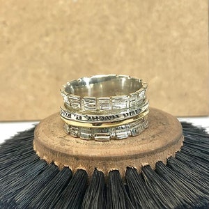 May include: A silver and gold spinning ring with a textured design. The ring is on a wooden base with black bristles.