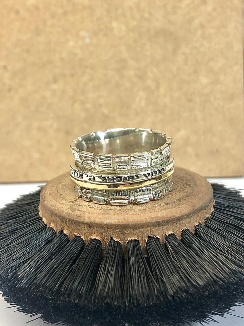 May include: A silver and gold spinning ring with a textured design. The ring is on a wooden base with black bristles.