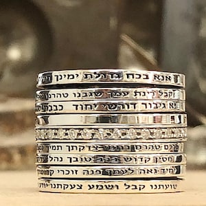 Puede incluir: Una pila de anillos de plata con texto hebreo grabado en cada banda. Los anillos están apilados uno encima del otro, siendo el anillo superior el más grande y el anillo inferior el más pequeño. Los anillos son todos de plata y tienen un acabado pulido.