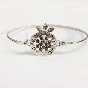 May include: A silver bangle bracelet with a pomegranate charm. The charm is decorated with red gemstones and Hebrew writing.