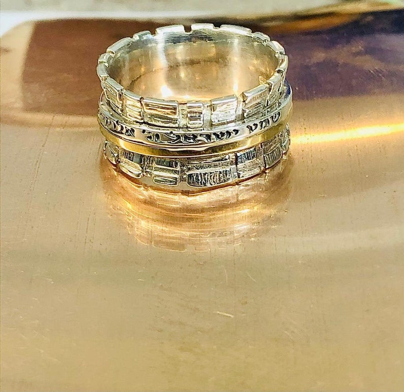 May include: A silver and gold spinning ring with a textured design. The ring has a band of Hebrew text.