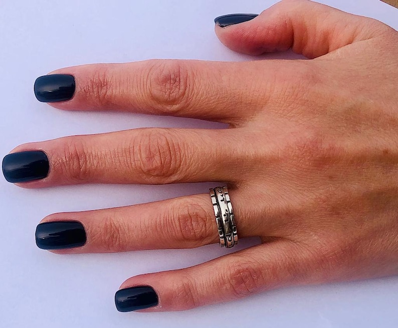 May include: A hand with black nail polish is wearing a silver ring with a textured design. The ring is made of three bands stacked together.