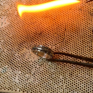 May include: A silver ring with Hebrew lettering is being heated with a torch on a perforated ceramic surface.