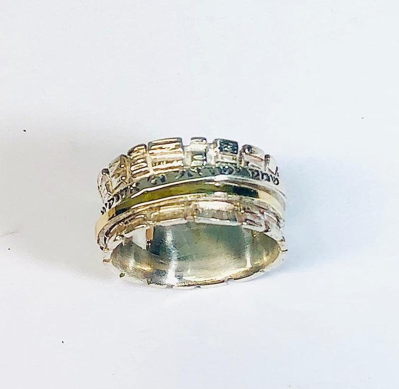 May include: A silver and gold ring with a textured design. The ring has a band of gold around the middle and is inscribed with Hebrew text.
