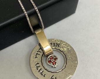 May include: A silver necklace with a round pendant featuring a pomegranate design with red gemstones. The pendant has Hebrew writing around the edge. The necklace has a box chain and a gold clasp.