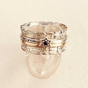 Puede incluir: Un conjunto de tres anillos de plata apilados uno encima del otro. El anillo superior tiene un diseño texturizado y una estrella negra con una pequeña piedra negra en el centro. El anillo del medio es una banda delgada con un acabado dorado y letras hebreas. El anillo inferior es una banda de plata texturizada.