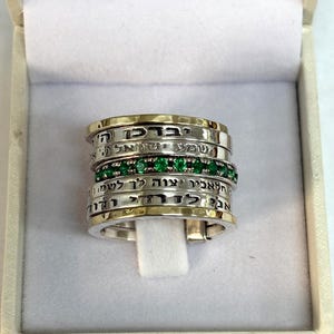 May include: A silver and gold ring with Hebrew text. The ring has multiple bands, some with gold rims and others with a row of green gemstones. The ring is displayed in a white box.