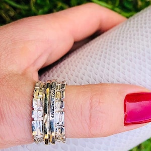 May include: A set of four silver and gold stacking rings. The rings are made of textured metal and have a geometric design. The rings are worn on a finger with red nail polish.