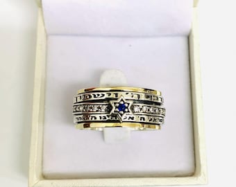 May include: A silver and gold ring featuring a Star of David with a blue center stone. The ring has multiple bands with Hebrew text and small clear stones. The ring is presented in a white box.