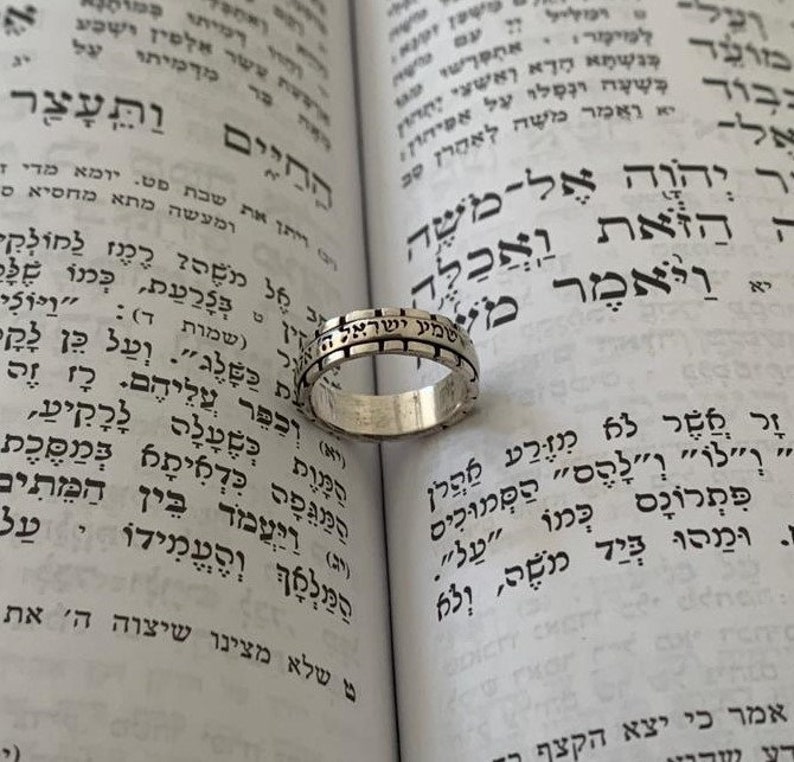 May include: A silver ring with Hebrew text inscribed around the band. The ring is resting on an open book with Hebrew text.