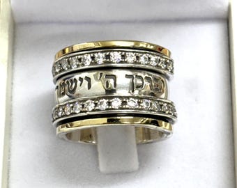 May include: A silver and gold ring with a spinning band. The ring features a gold band on the top and bottom, with a silver band in the middle. The silver band has Hebrew text and is encrusted with small, clear stones. The ring is in a white box.