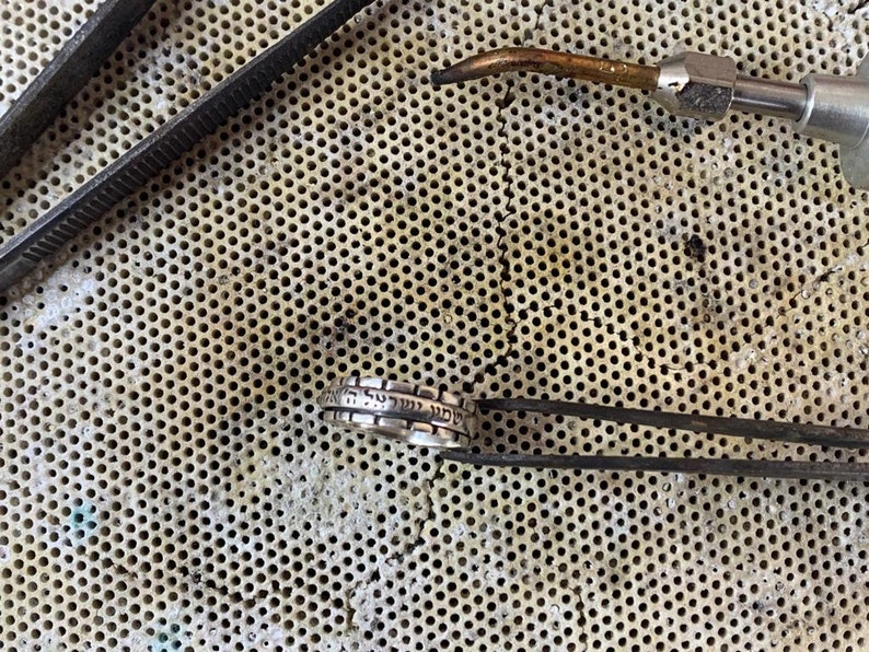May include: A silver ring with Hebrew lettering on a metal workbench with tools.