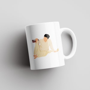 May include: White ceramic mug with a graphic of a person sitting and pouring a liquid from a pitcher. The person is wearing a white outfit and has dark hair.
