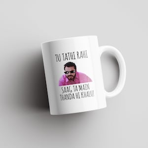 Könnte beinhalten: Weiße Keramiktasse mit einem Schwarzweißbild eines Mannes mit Sonnenbrille und rosafarbenem Hemd. Der Text "Tu tathi rahi saag ta main thanda hi khalu!" ist auf die Tasse gedruckt.
